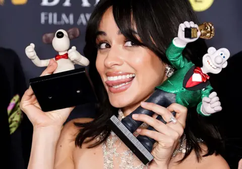 PA Media Camila Cabello sticks her tongue out at the camera as she holds up models of Wallace and Gromit