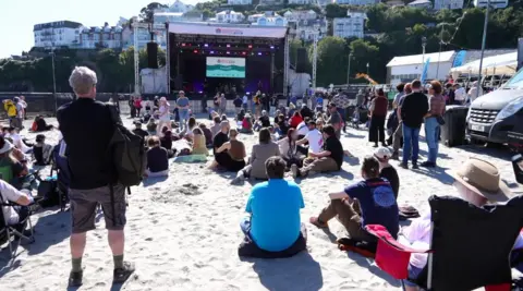 A large festival stage on a beach. Scores of people are sitting in front of it. 