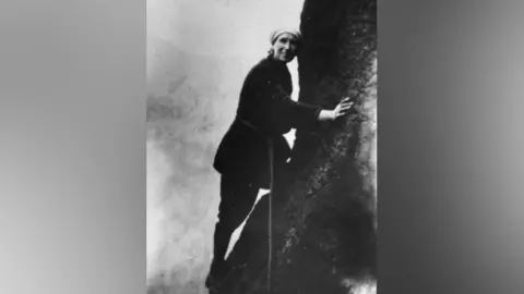 A black and white photograph (in the early days of cameras) shows Mable Barker wearing a white cap and thick dark-coloured clothing while climbing Girdle Traverse in the Lake District. She has a rope tied around her waist. 