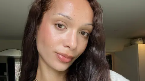 Billie van der Westhuizen, 30, with long dark wavy hair and a white jumper taking a selfie with a neutral facial expression