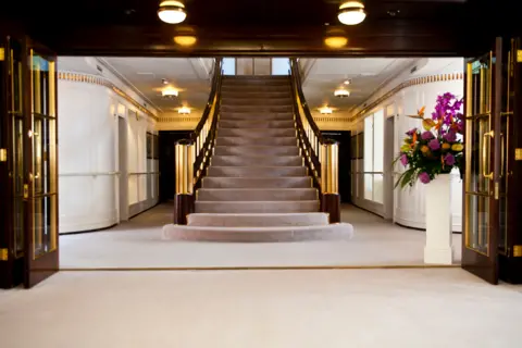 Marc Millar I stair case in the middle of the room. The room has white carpet and walls with brown wood and gold detailing.