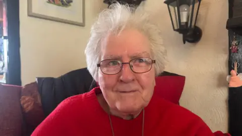 An elderly woman with short grey hair, glasses and a red jumper is sat on a burgandy sofa in what appears to be a living room