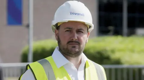 Patrick McIntyre - a man with a thin beard and moustache, with dark hair. He has a high vis jacket and white hard hat on. 