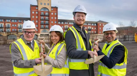 Richard Parker, Mayor of the West Midlands, Maggie Grogan, Muse - Managing Director Midlands, Councillor Chris Burden, City of Wolverhampton Council, and Basit Ali, Muse Development Director, at the Smithgate phase one site. They are wearing hard hats, yellow-hi vis vests and holding two spades between them. The ground is mud, with a building in the background.