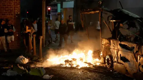 Property is set on fire in a street at night. A crowd, obscured by shadow and with their backs to the camera, watch on.