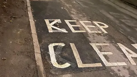 A stretch of newly-patched road with the words "Keep Clear" repainted on top of the K and C the incorrect way around