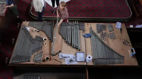Around one hundred pipes which have been removed from the pipe organ lay on a large table in front of the organ. Volunteers dust and clean them with paint brushes and cleaning products. 