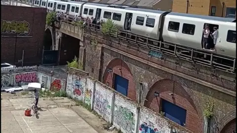 A delivery rider stands in a graffiti-covered yard below an elevated train track, preparing to throw a bottle of water to stranded passengers leaning out of a stopped train in the heat.