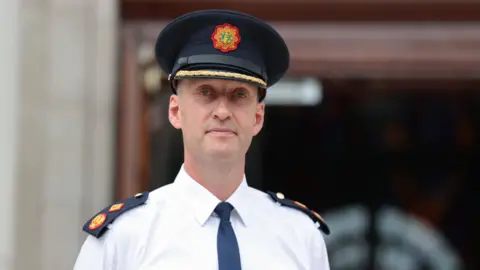 PA Media A man standing outside wearing a police uniform consisting of a hat, shirt and tie. The entrance to a building can be seen out of focus behind him.