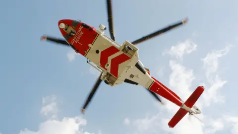 A view from the ground up towards the underside of a coastguard helicopter. It is viewed against a blue sky with clouds. The helicopter has red and white markings.