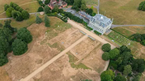 An aerial view of Kingston Lacy showing large areas of brown, drought-affected grass surrounding the historic house, with pathways crossing the grounds and greener trees around the edges of the estate.