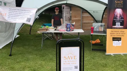 Save Novello Picture House A stand at a fair with signs and posters about the Novello Theatre campaign. A person is standing under a gazebo behind a table laid out with bags and other items.