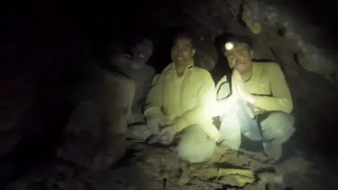 A group of men crouch inside a dark cave as they are being rescued.