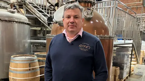 Leanne Rinne/BBC The co-founder of the Copper Rivet Distillery in Chatham, Matthew Russell, pictured in the distillery. In the background there are several copper pots for making the gin and several barrels for storing it. Matthew has greying hair and is wearing a navy branded jumper and pink undershirt.