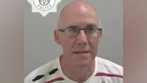 West Mercia Police A mugshot of a bald man wearing black glasses, and a white t-shirt with a red and black horizontal stripe.