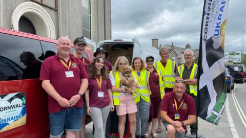 Ten people stood together, They are all looking at the camera and smiling. Some of them are wearing maroon tops and others are wearing green high visibility clothing. There is a red van and a white van behind him. There is a banner to the right.
