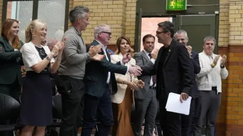PA Media A group of men and women clap as they line up to greet a dark-haired man in a black suit, black jumper, and black glasses. who shakes hands with a man with white hair and dark framed glasses in a navy suit and white shirt. The people are standing by a green doorway in a building, the door ajar on to a street.