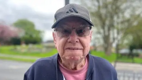Man staring into the camera by the roadside in Waltham, North East Lincolnshire. He is wearing glasses, a grey sports cap along with a pink t-shirt and blue casual jacket. Across the road behind him are trees and a flower bed. 