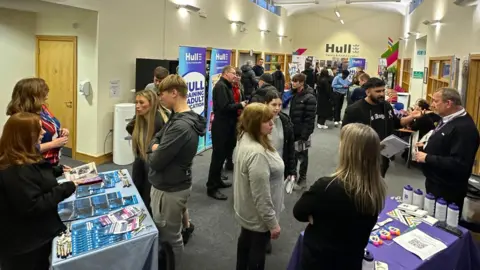 A crowd of people gathered at an event inside a large room. There are various displays and stands and banners which say Hull Training and Adult Education on them.