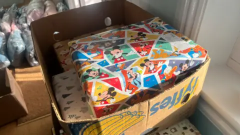 A box of wrapped presents. The top parcel has Micky Mouse wrapping paper on it which is colourful. Below it is white wrapping paper with green Christmas trees on it.