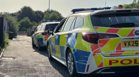 Charlie Stubbs / BBC There is two police cars parked up on a potholed narrow road. On the left hand side, there is metal gates, some dark brown fences and some weeds. In the background are some trees and a lamp-post.