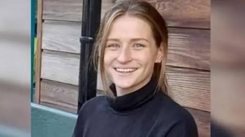 A young woman with long hair in a pony tail and wearing a black turtle neck smiles at the camera. She is sitting in front of a shed.