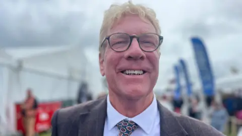Kim Conchie of the Cornwall Chamber of Commerce smiles while stood at the Royal Cornwall Show. He has a suit on with a tie and is wearing glasses.