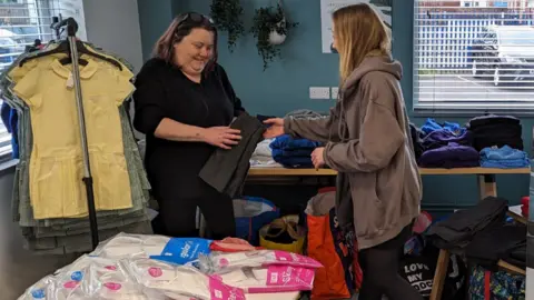 Little Stars Charity A woman in a black t-shirt is holding some grey children's trousers. A woman to her right, wearing a brown hoodie, is passing the trousers to her. To the left is a clothes rail with coloured checkerboard school dresses hanging on it. There are also tables piled with other clothing.