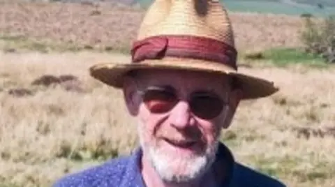 Police Scotland A man with a grey beard is wearing blue shirt, a straw hat and dark glasses. There is grassland behind him