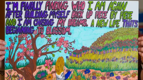 Peterborough City Council A posted of a painting shows a woman sitting in a field looking at a grass hill with colourful flowers on it. Behind her is a tree with pink flowers blooming on it. Words on the poster read: "I'm finally finding who I am again after building myself back up piece by piece and I am chasing my dreams. A new life that's beginning to blossom."