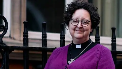 A woman standing on the pavement, she has black curly hair and is smiling while wearing a purple cardigan 