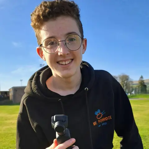 Edward Ryan A young man with curly hair and glasses holding a small camera