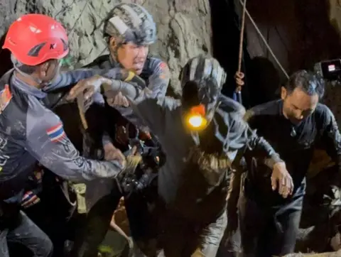 Two rescuers in dark blue diving gear, with one wearing a black helmet and the other a red one, hold the right arm of the rescued man, who is dark muddy clothes and has a headlight on. His left arm is being held by another rescuer to his left as they all emerge from the cave in darkness