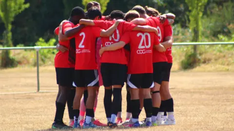 A group of players in a huddle, on a football pitch. They are wearing red tops and black shorts. Foliage is behind them. 
