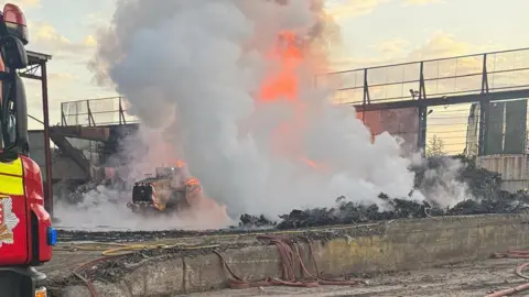 Essex Fire and Rescue The same scene in daylight, showing charred metal on the ground and smoke and fire continuing to billow from the scene. A vehicle appears to be in the midst of it, presumably also on fire.