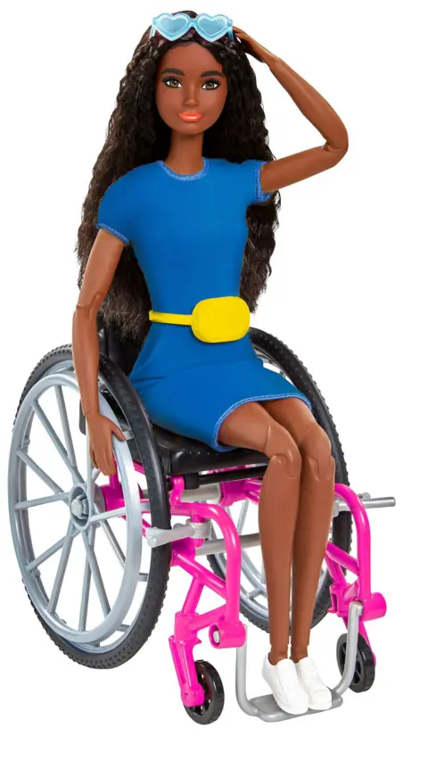 Mattell Inc Fashion doll seated in a pink-framed wheelchair with large grey wheels, wearing a blue dress with a yellow belt, white sneakers, and heart-shaped blue sunglasses on the head, against a plain light background.