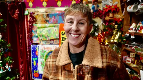 Lisa is wearing a checked coat and standing in her stall, surrounded by bright Christmas ornaments and wrapping paper.