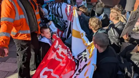 A group of people holding banners sit on a pavement
