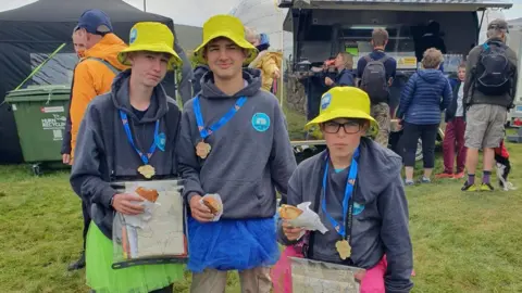 A group of three children wearing yellow hats are standing next to each other eating rolls. They are smiling. Behind them is a food stall.
