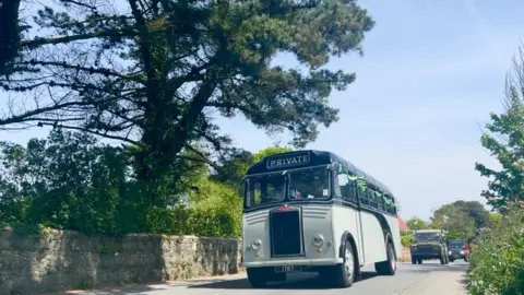 A vintage grey and black bus driving down a street in guernsey as part of the cavalcade
