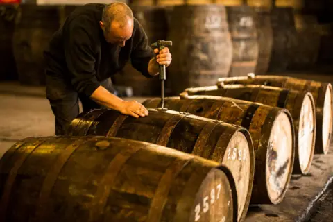 Getty Images A man in a black jumper and dark trousers bent over a row of whisky casks in a distillery. He is opening one of the barrels with a metal tool.
