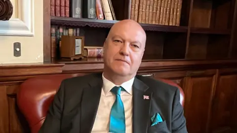 Ian Cooper sits in a chair in the library of the county building. He is wearing a suit and blue tie, with a union flag badge pinned to his jacket. 