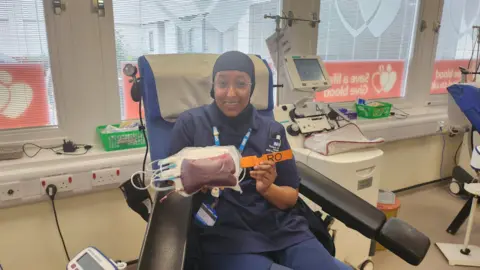 Amino Ali is wearing glasses, a head covering and a dark blue nurse uniform. She is smiling and sat in a blood donation chair. She holds up a bag of blood with an Ro label attached to it. 