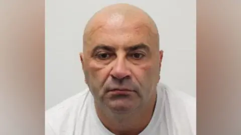 Mugshot of Bald man in white shirt looks below eyeline of camera