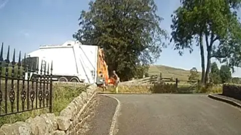 Dave Fishwick A grainy image of a road. On the left side, a small knee-high wall and black metal fence. Straight ahead, a white bin wagon, with a man carrying a bin behind it. In the distance trees, a green hill and a blue sky can be seen