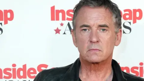 A man wearing a leather jacket and a black top standing in front of the press,
there is a backdrop behind him that reads inside soap awards 2024