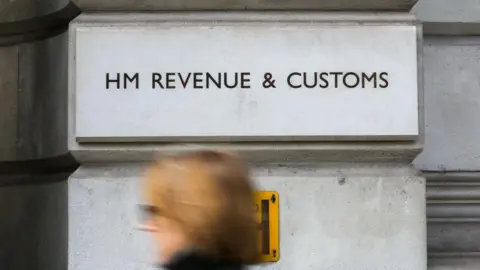 The HM Revenue & Customs logo on a pale brick. A woman walks past in the foreground.