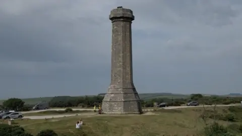 A grey stone tower standing in the country side. It towers over the cars parked nearby with four people standing around the base. The tower is surrounded by grassland with trees dotted in the landscape.