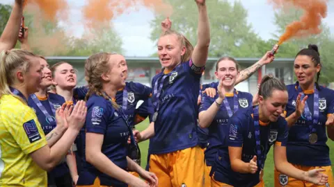 Hull City Ladies football team celebrating on the field, wearing navy and amber kits. One player is raising her arm as others are cheering and smiling. Players have blue medals around their necks. Orange smoke flares fill the air. Green trees can be seen in the background. 
