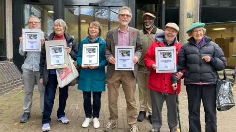 Seven people, three men and four women are holding signs and smiling outside a council building.